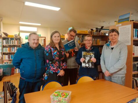 Świetlicy Terapeutycznej dwóch mężczyzn i dwie kobiety. kobieta i mężczyzna trzyma książkę, w tle regały z książkami. Zdjęcie we wnętrzu biblioteki.