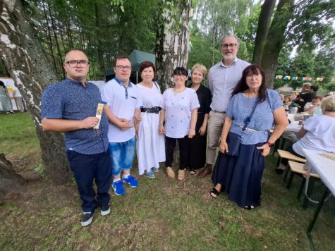 Na zdjęciu stoja kobiety i męzczyżni, uczestnicy Świetlicy Terapeutycznej wraz z opiekunami. W tle drzewa i trawa.
