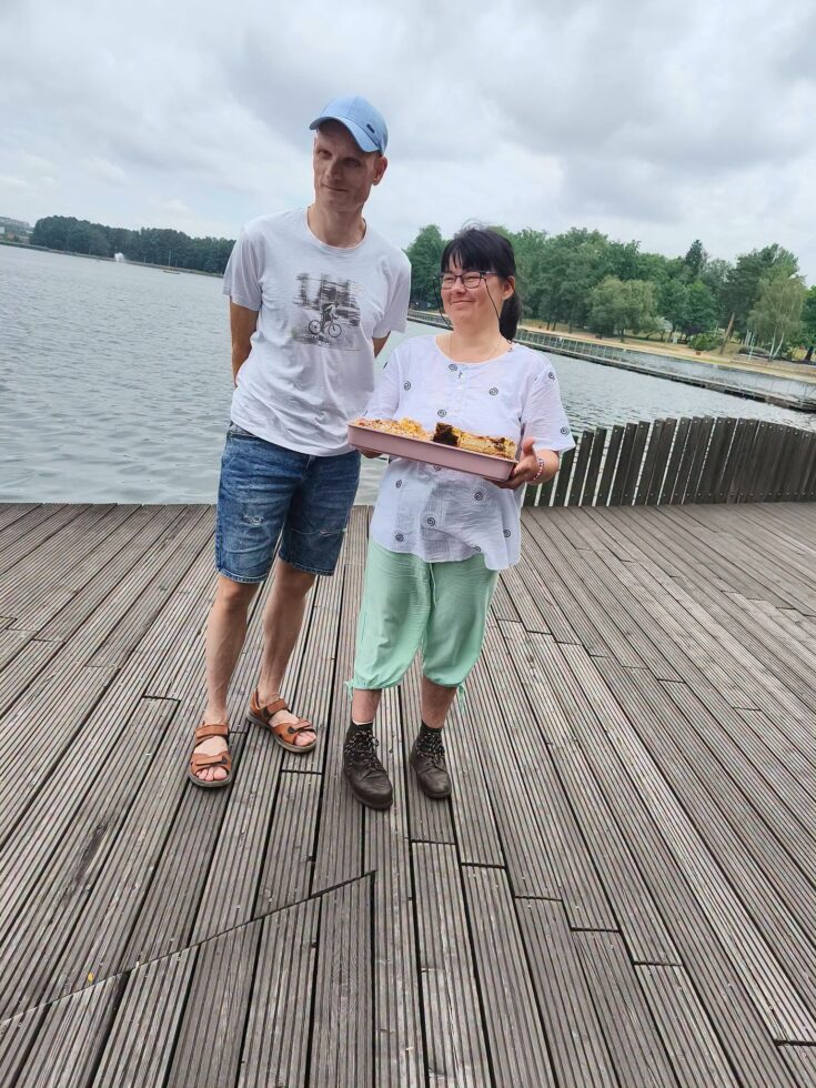 Na zdjęciu na pomoście stoi kobieta w okularach trzyma tacę z ciastem, obok niej stoi mężczyzna w czapce z daszkiem. W tle jezioro i plaża oraz drzewa.